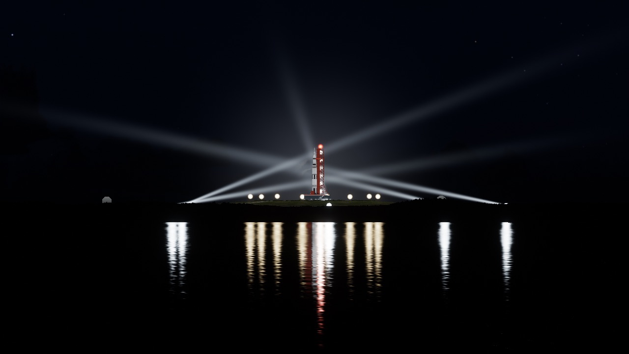 The rocket on its pad before dawn, lit by searchlight beams, its lights reflected in the water.